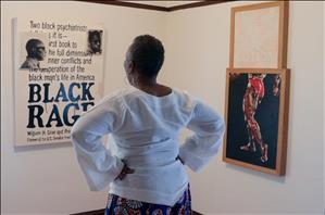 A Black woman with her hands on her hips looks at artworks. Her back is to the camera. 